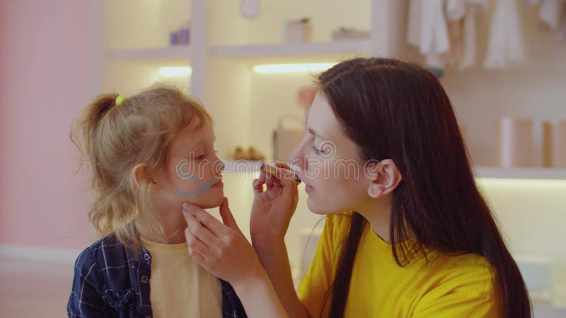 Portrait of positive mom face painting cat whiskers on little girl face