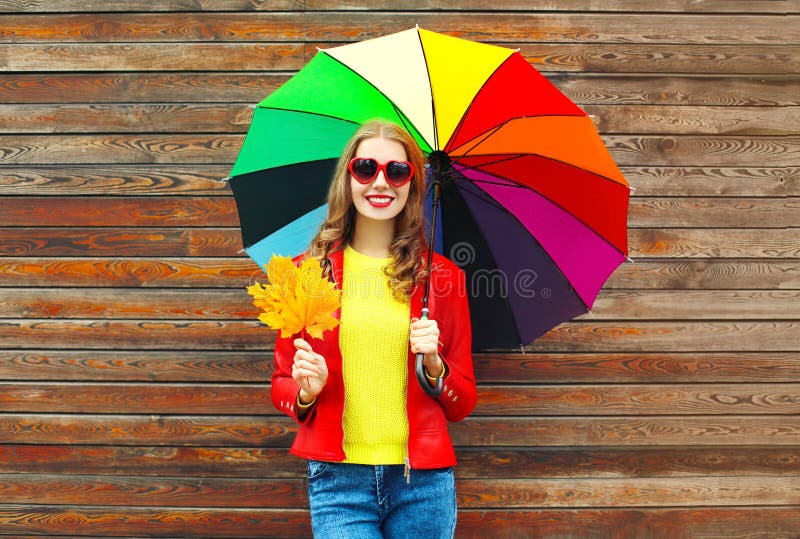 Portrait pretty smiling woman with colorful umbrella in autumn with maple leafs over wooden background wearing a red leather jacket