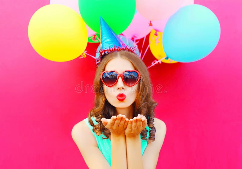 Portrait pretty woman in a birthday cap is sends an air kiss holds an air colorful balloons on pink background