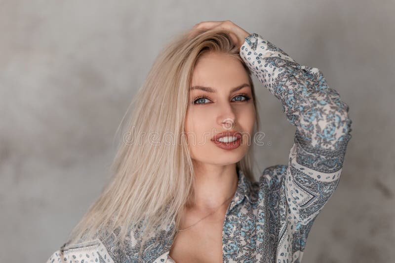 Portrait of a pretty young blonde woman with blue eyes with plump sexy lips in a fashionable gray summer shirt with a pattern in a studio near the wall. Beautiful european girl. Female face portrait