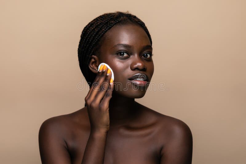 Portrait rapproché d'une femme afro-américaine séduisante qui enlève du maquillage avec une éponge isolée sur fond beige photo stock