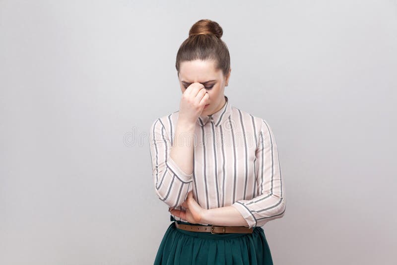 Portrait of sad depressed young woman in striped shirt and green skirt with collected ban hairstyle, standing, holding head hown