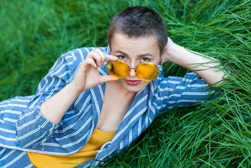 Portrait of serious cute young woman with short hair in casual blue striped suit, yellow shirt and eyeglasses lying down on green