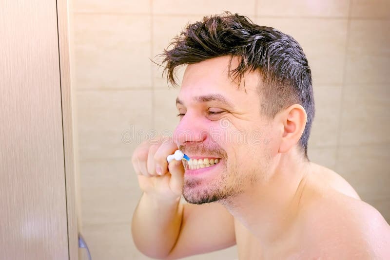 Shaggy sleepy young man intensively cleaning teeth in bathroom, side view.