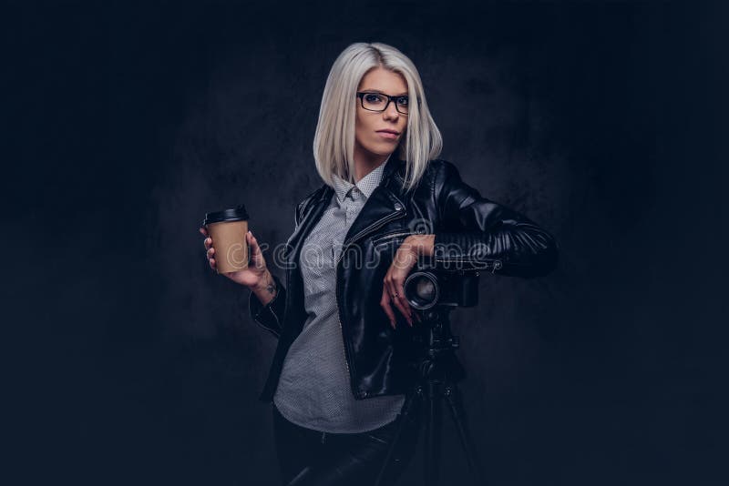 Portrait of a smart blonde female photographer in trendy clothes and glasses holds a takeaway coffee and posing while leaning on a professional camera with a tripod at a studio, looks at the camera. Isolated on a dark background.