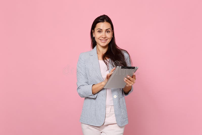 Portrait of smiling charming young woman in striped jacket using tablet pc computer isolated on pink pastel wall