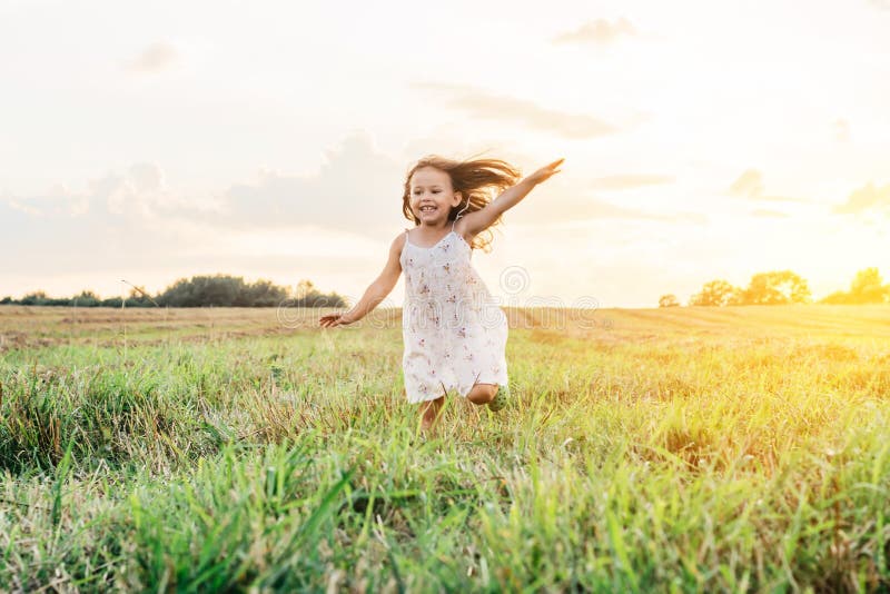 Portrait of smiling girl playing, jumping and running on grass hay meadow paths of dry grass in the sunset. Bright sky