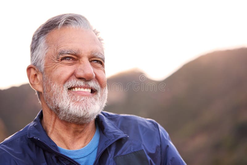 Portrait Of Smiling Hispanic Senior Man Outdoors In Countryside With Mountains In Background