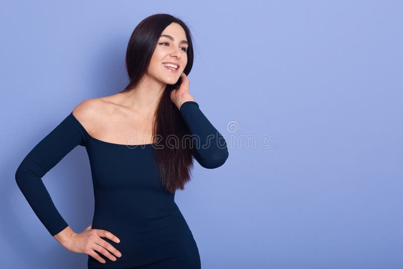 Portrait of smiling pretty young woman wearing dress with bare shoulders, looking aside isolated blue background in studio. People