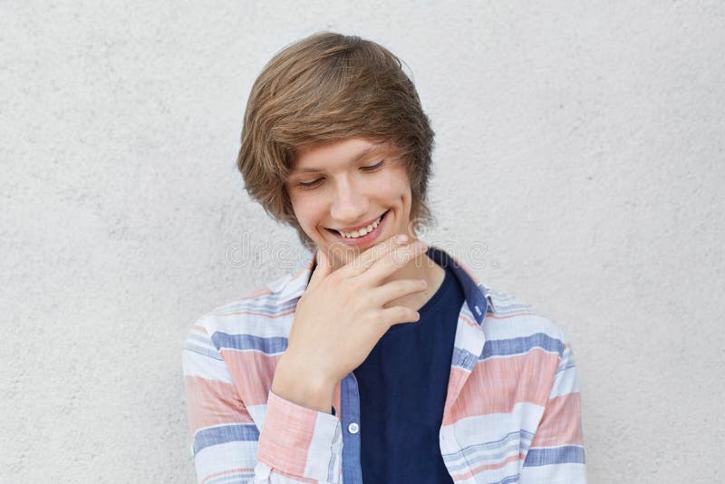 Portrait of smiling shy boy with trendy hairdo wearing casual shirt looking down holding hand on chin having dimple on his cheek.