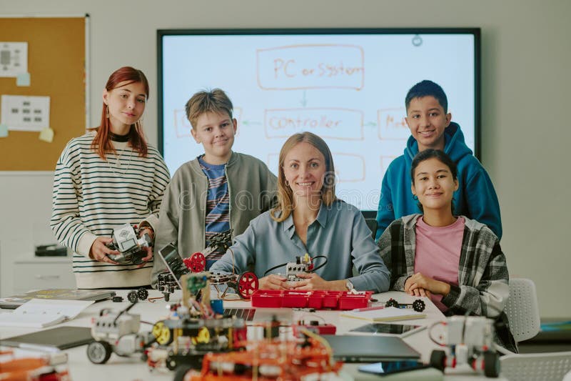 Portrait of Teacher and Students Together in Classroom. Portrait of smiling teacher sitting at table and surrounded by multiracial students in classroom of robotics with huge number of robots and its parts