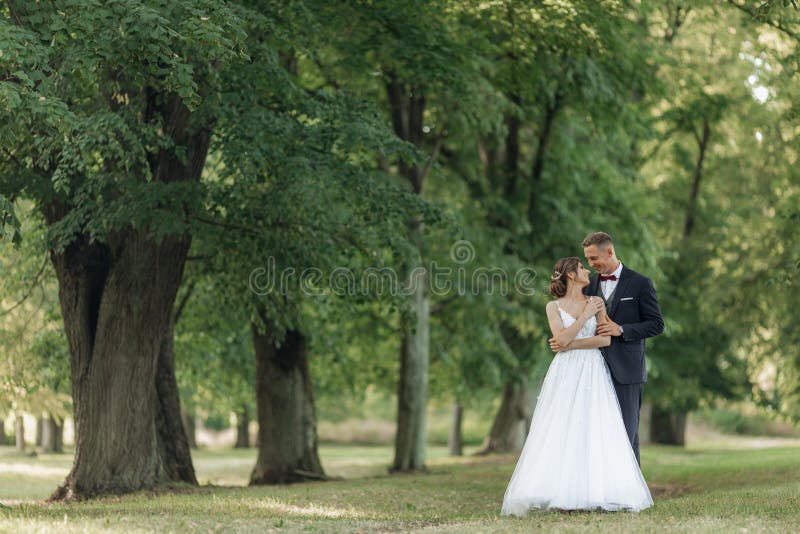 Portrait of smiling wedding couple stand on green grass in park in summer. Young beautiful bride looking at young man.