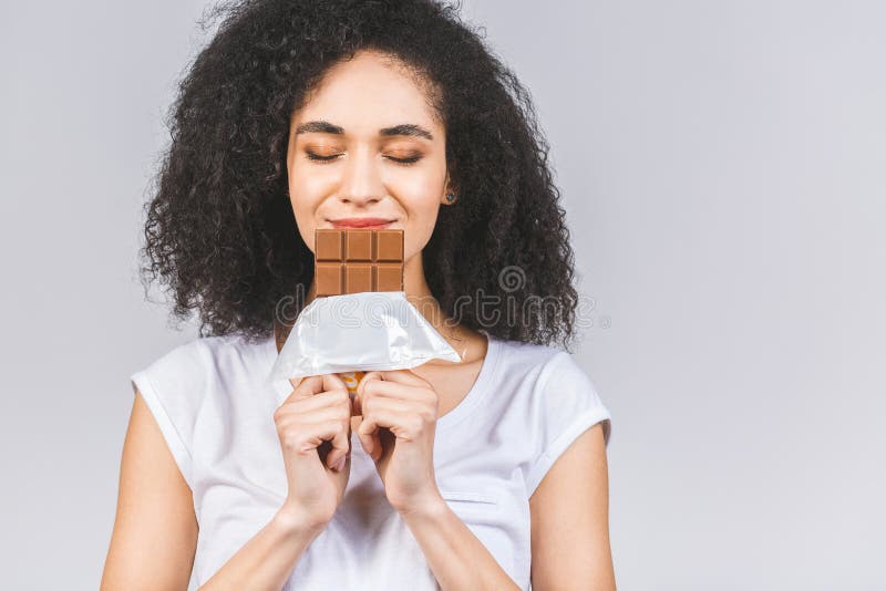 Portrait of a smiling young african american black woman eating chocolate bar isolated over grey background