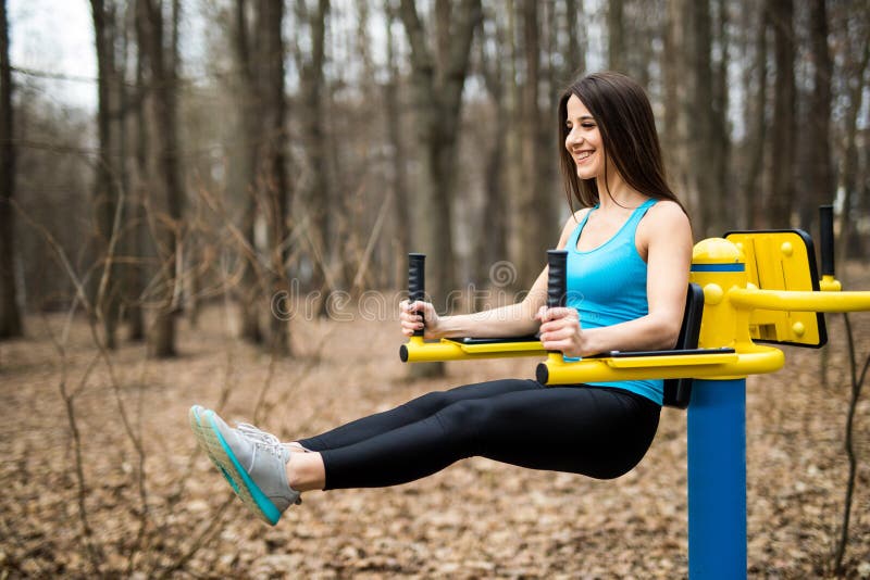 Portrait of strong young woman hanging on wall bars with her legs up. Fitness woman performing hanging leg raises on outdoor in pa