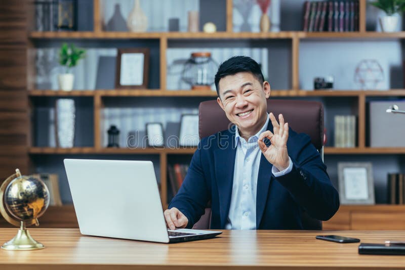 Portrait of a successful Asian businessman, in a classic office, the boss looks at the camera and smiles, shows a sign ok hand