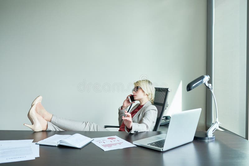 Portrait of successful female boss sitting at big desk in modern office speaking by phone with feet on table. Portrait of successful female boss sitting at big desk in modern office speaking by phone with feet on table