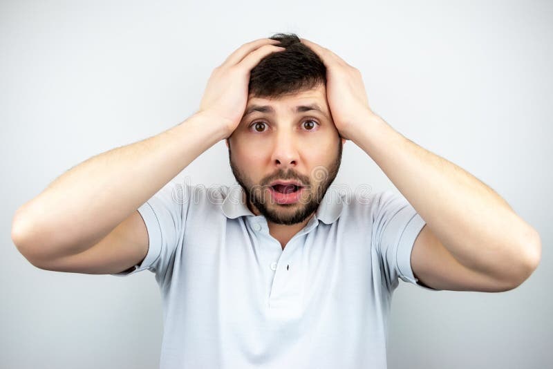 Portrait of a surprised bearded man with eyes and mouth wide open, holding his head with his hands, in a white t-shirt on a white background. Portrait of a surprised bearded man with eyes and mouth wide open, holding his head with his hands, in a white t-shirt on a white background