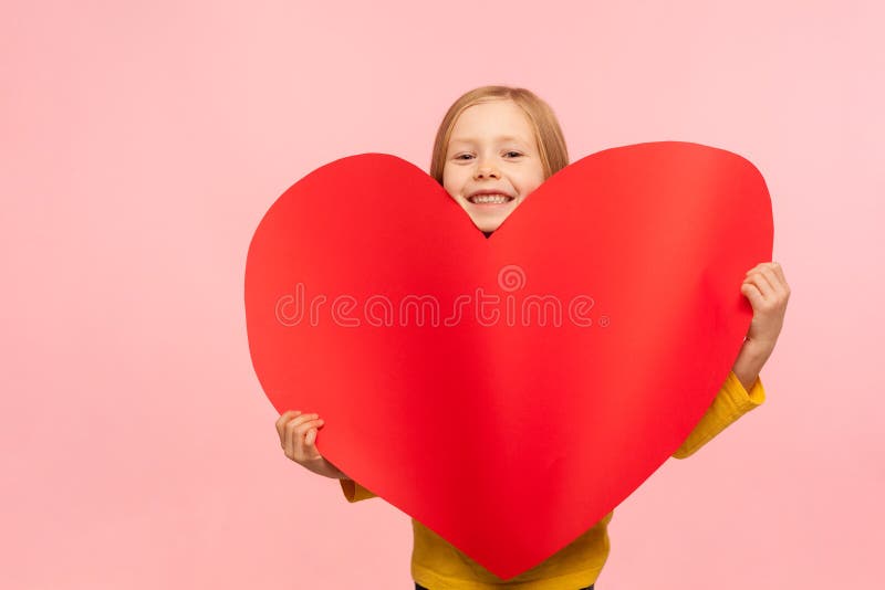 Portrait of sweet adorable little girl peeking out of large red heart symbol and smiling to camera, congratulating on Mother`s day, saying I love you. indoor studio shot isolated on pink background