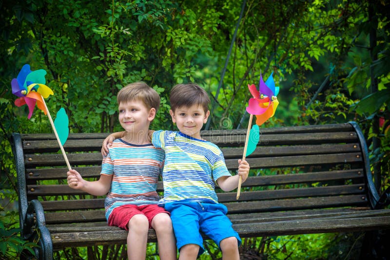 Portrait of two boys, sibling brothers and best friends smiling. Kids sitting on bench play together with pinwheel. Outdoors royalty free stock photo Portrait of two boys, sibling brothers and best friends smiling. Kids sitting on bench play together with pinwheel. Outdoors royalty free stock photo