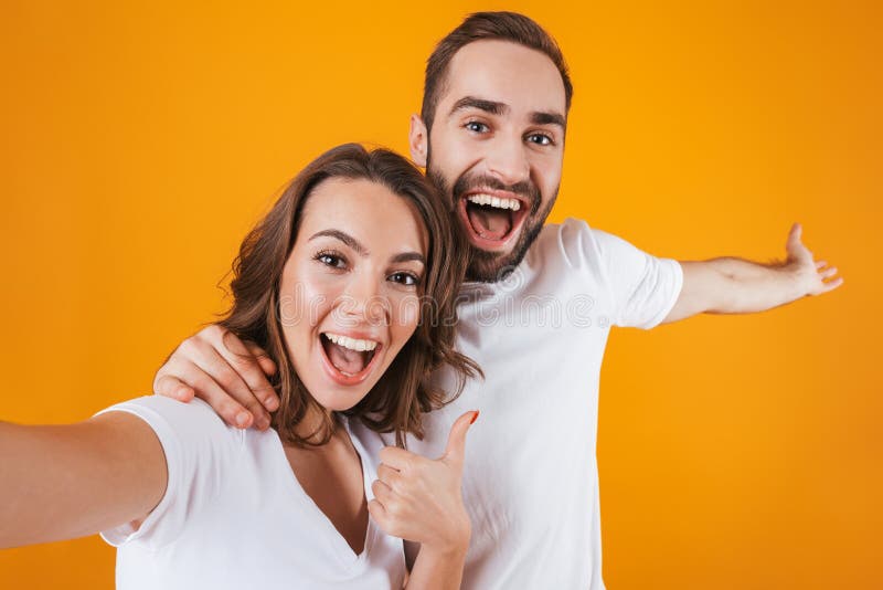 Portrait of two happy people man and woman smiling while taking selfie photo, isolated over yellow background