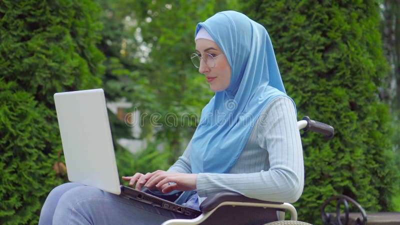 Portrait une jeune femme arabe handicapÃ©e dans une Ã©charpe traditionnelle utilise un ordinateur portable assis dans un fauteuil banque de vidéos