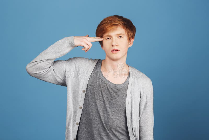 Portrait of unhappy attractive ginger male student in casual gray outfit holding hand in gun gesture near hand, being stock photo Portrait of unhappy attractive ginger male student in casual gray outfit holding hand in gun gesture near hand, being stock photo