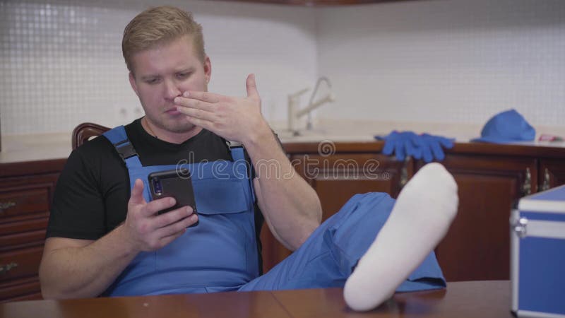 Portrait of unscrupulous caucasian repairman sitting at kitchen with legs on the table and stretching foot.