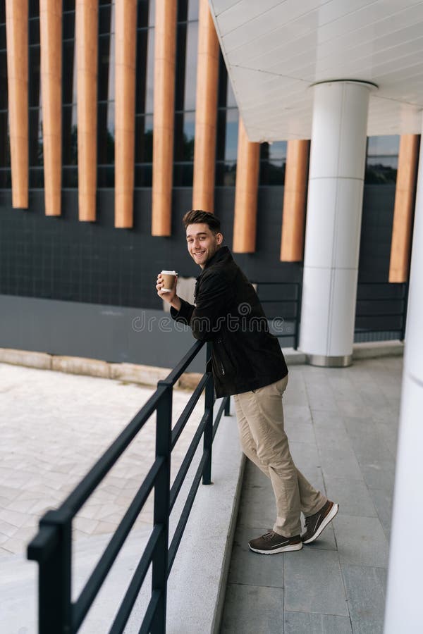 Portrait vertical complet d'un jeune homme heureux tenant une tasse à café à emporter sur une rampe debout par un moderne images stock