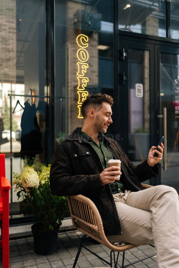 Portrait vertical d'un jeune homme heureux assis sur une chaise de café en plein air tenant une tasse de café à emporter et navigu photos libres de droits