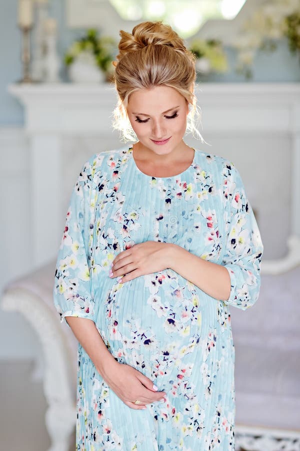 The portrait is very beautiful, mile, feminine and tender pregnant girl blonde in blue dress with floral print against a light in stock images The portrait is very beautiful, mile, feminine and tender pregnant girl blonde in blue dress with floral print against a light in stock images