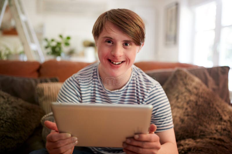 Portrait Of Young Downs Syndrome Man Sitting On Sofa Using Digital Tablet At Home. Portrait Of Young Downs Syndrome Man Sitting On Sofa Using Digital Tablet At Home