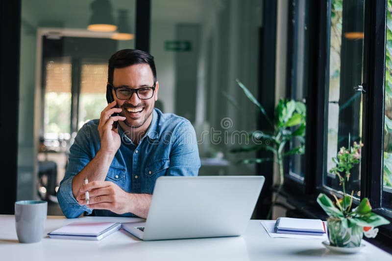 Portrait von jungen, fröhlichen Unternehmern in Gelegenheitsbüros, die Telefongespräche führen, während sie mit Laptop arbeiten stockbilder