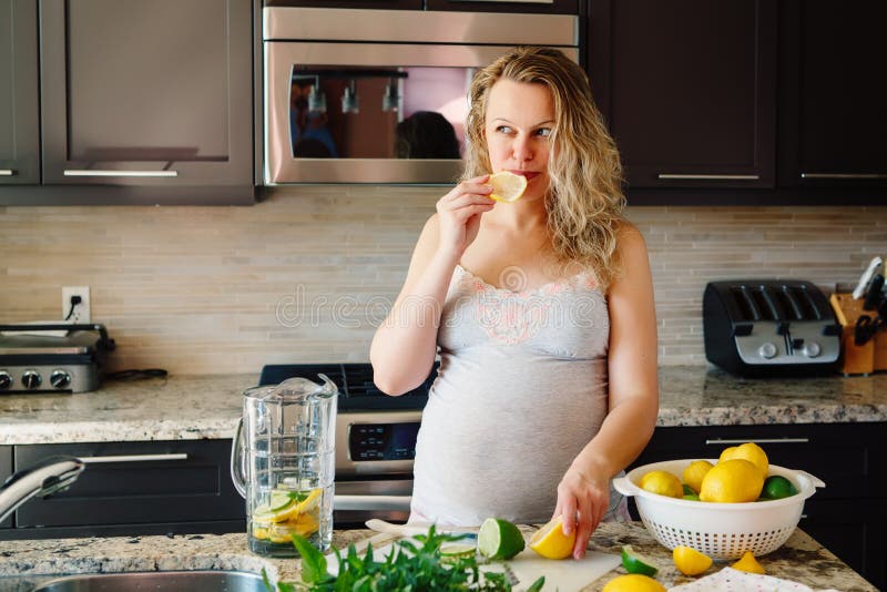 Portrait of white Caucasian blonde pregnant woman eating citrus lemon making juice standing in kitchen at home looking away stock photography Portrait of white Caucasian blonde pregnant woman eating citrus lemon making juice standing in kitchen at home looking away stock photography