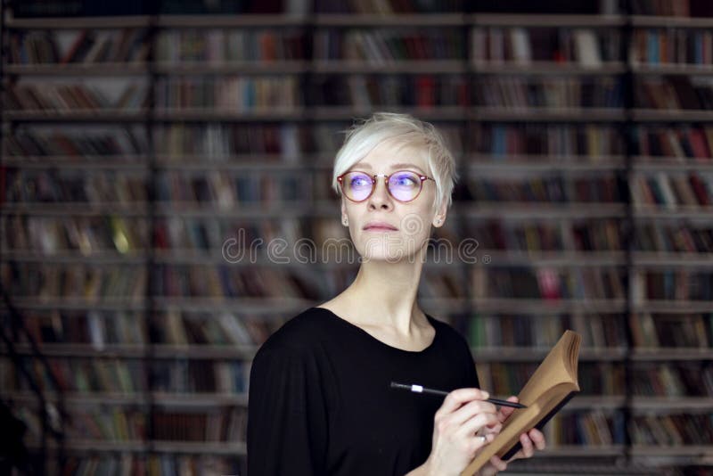 Portrait of woman with blonde hair and eyeglasses in a library, opened book. Hipster student. Education concept stock photo Portrait of woman with blonde hair and eyeglasses in a library, opened book. Hipster student. Education concept stock photo