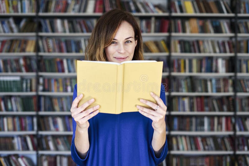 Portrait of woman in a library face half-covered by opened book, reading. College student in co-working, concept of studying