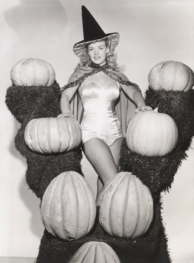 Portrait of woman in witch costume surrounded by pumpkins on grass shelves