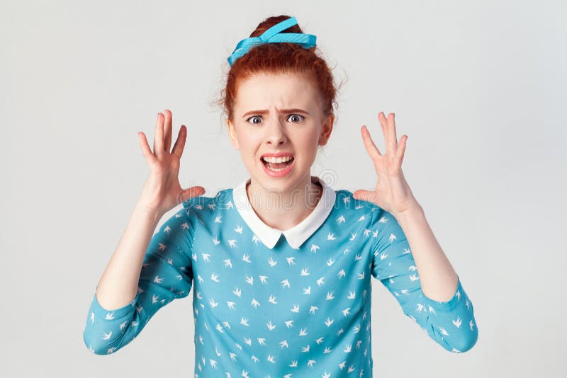 Girl in despair and shock. Portrait of young angry redhead girl in blue dress looking panic, screaming in camera with mouth wide open, with hands up. studio shot on gray background.