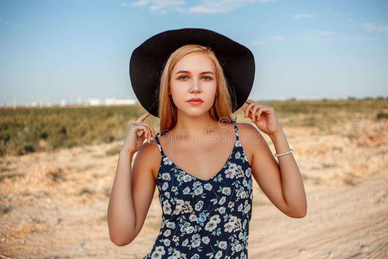 Portrait of a young beautiful caucasian blonde girl in a blue dress with a floral print and a black hat standing on a sandy stock photography Portrait of a young beautiful caucasian blonde girl in a blue dress with a floral print and a black hat standing on a sandy stock photography