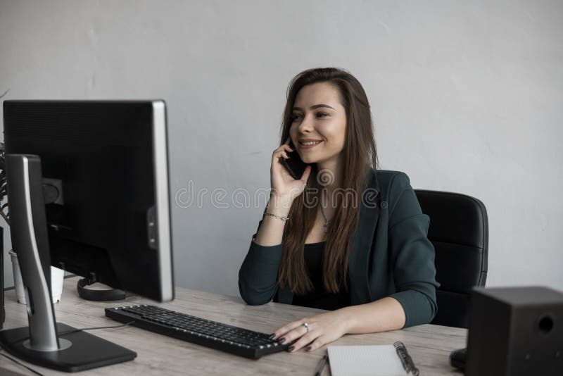 Portrait of young business woman talking phone against computer screen in white office. Female entrepreneur having call