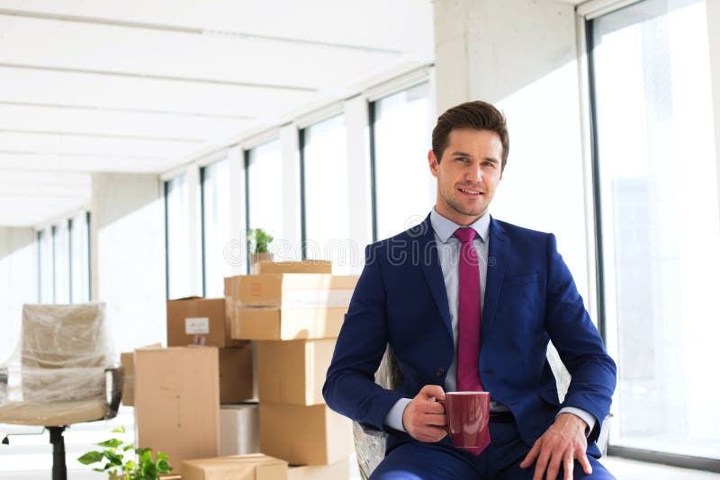 Portrait of young businessman having coffee with moving boxes in background at office stock photography Portrait of young businessman having coffee with moving boxes in background at office stock photography