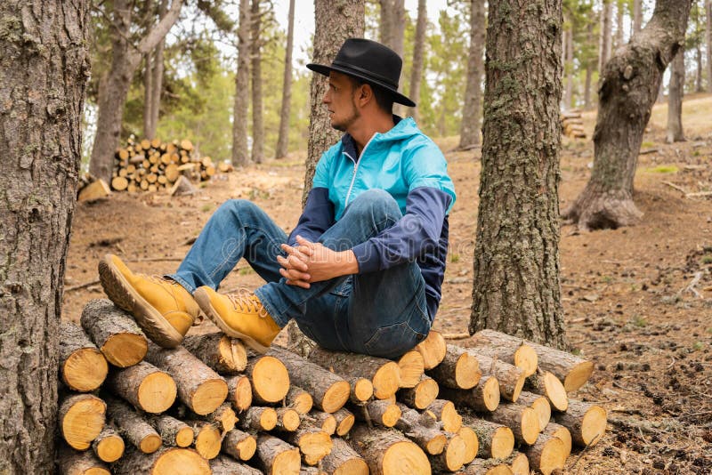 Portrait young caucasian man in black cowboy hat sitting on a pile of wooden logs in forest landscape.Male enjoying and resting in