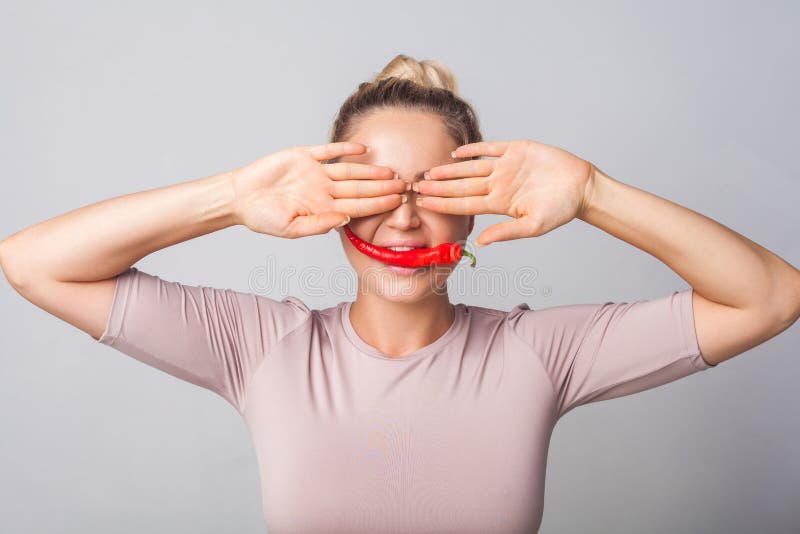 Portrait of young cute woman covering eyes with hands and biting red pepper, holding spicy chilli in mouth