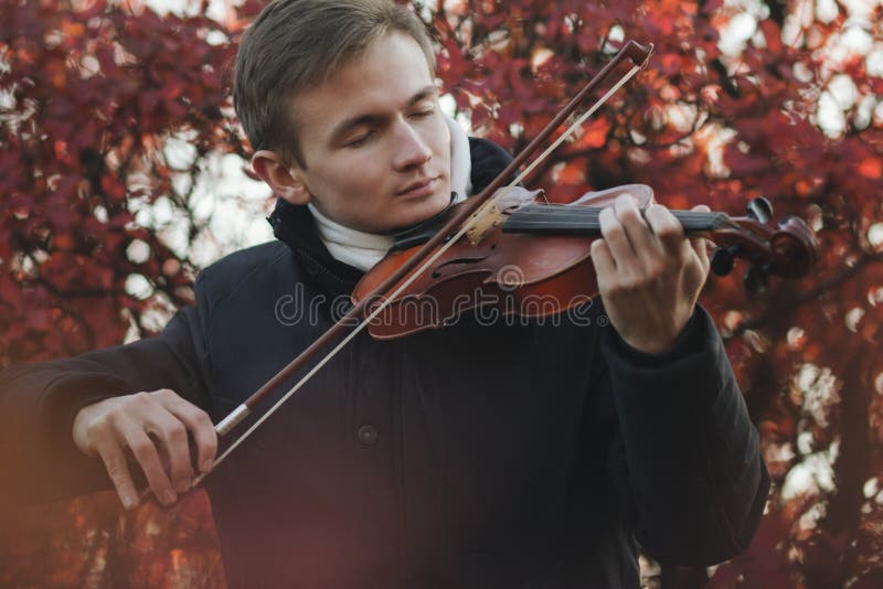 Portrait of a young elegant man playing the violin on autumn nature backgroung, a boy with a bowed orchestra instrument makes a concert, concept of classical music, hobby and art