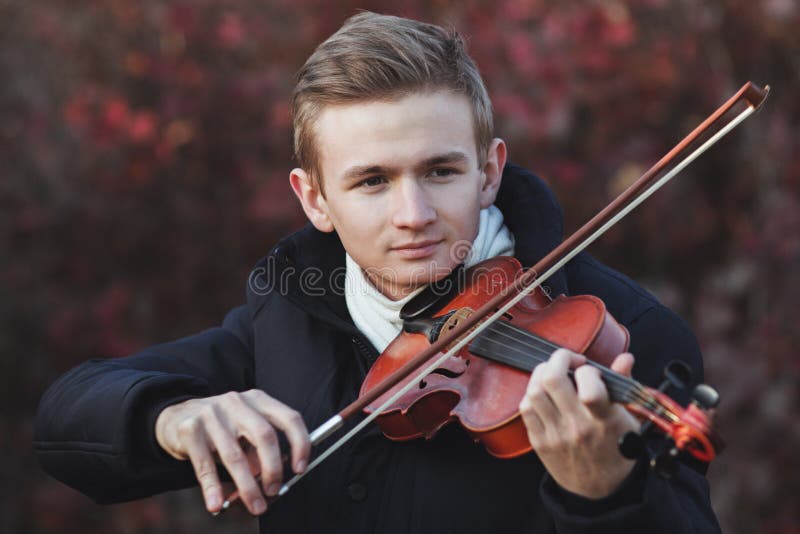 Portrait of a young elegant man playing the violin on autumn nature backgroung, a boy with a bowed orchestra instrument makes a concert, concept of classical music, hobby and art