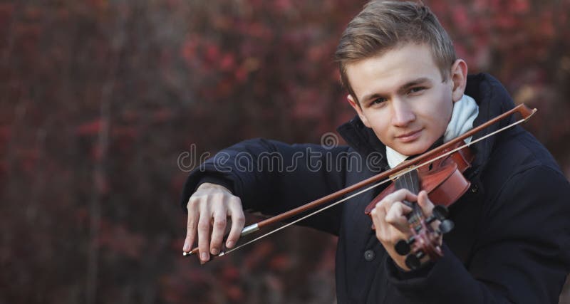 Portrait of a young elegant man playing the violin on autumn nature backgroung, a boy with a bowed orchestra instrument makes a concert, concept of classical music, hobby and art