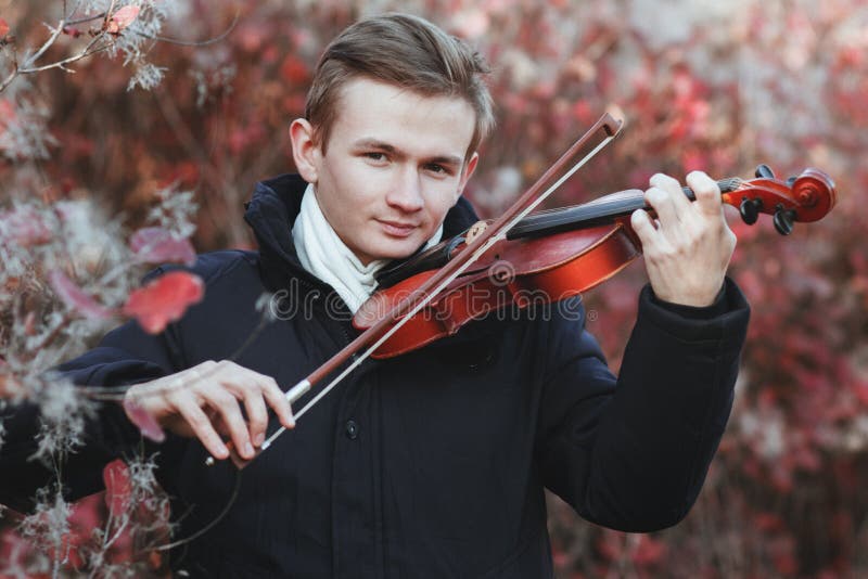 Portrait of a young elegant man playing the violin on autumn nature backgroung, a boy with a bowed orchestra instrument makes a concert, concept of classical music, hobby and art