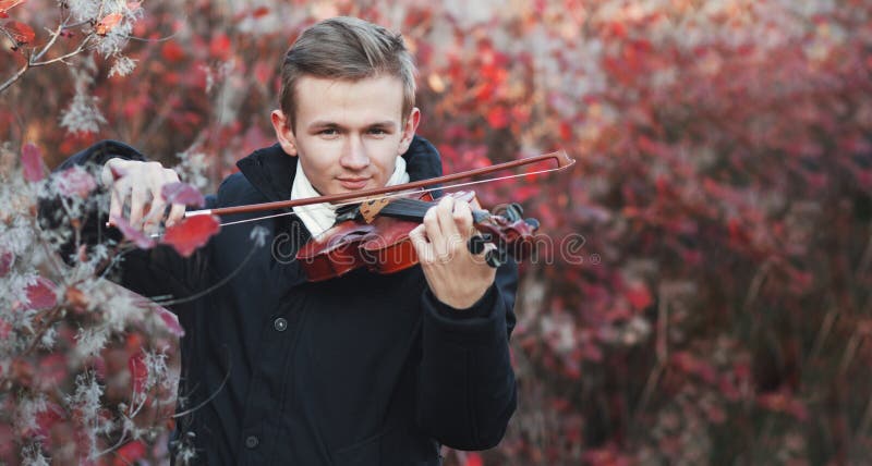 Portrait of a young elegant man playing the violin on autumn nature backgroung, a boy with a bowed orchestra instrument makes a concert, concept of classical music, hobby and art