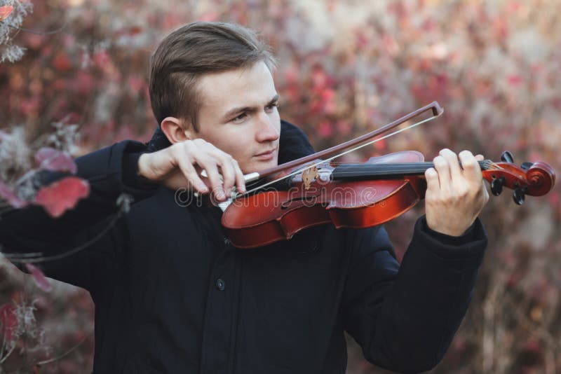 Portrait of a young elegant man playing the violin on autumn nature backgroung, a boy with a bowed orchestra instrument makes a concert, concept of classical music, hobby and art