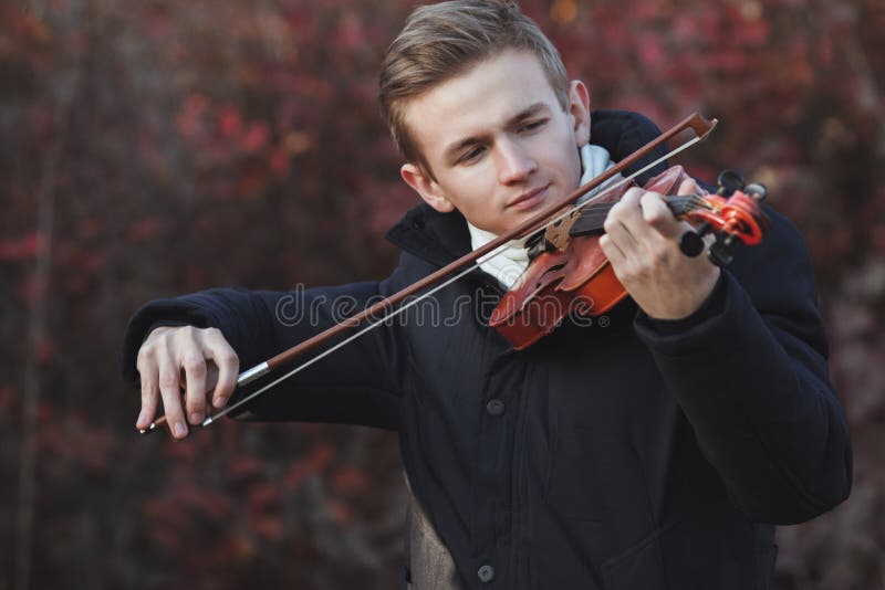 Portrait of a young elegant man playing the violin on autumn nature backgroung, a boy with a bowed orchestra instrument makes a concert, concept of classical music, hobby and art