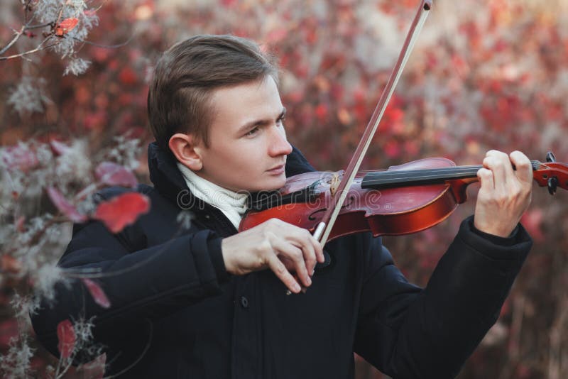 Portrait of a young elegant man playing the violin on autumn nature backgroung, a boy with a bowed orchestra instrument makes a concert, concept of classical music, hobby and art